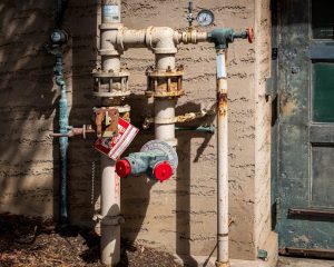Rusty outdoor plumbing pipes with pressure gauges and warning signs on a wall. | Wade's Plumbing & Septic